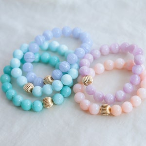 May include: Four beaded bracelets in pastel shades of blue, purple, and pink. Each bracelet has round, colourful beads and a gold-coloured, textured accent bead. The bracelets are displayed on a white, textured surface.