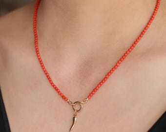May include: A necklace featuring a strand of small, round, vibrant orange-red beads. The necklace includes a gold-toned circular clasp at the centre, from which a single, slender, curved gold-toned horn-shaped pendant hangs downwards.