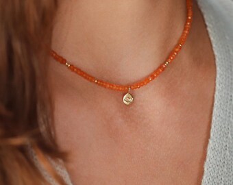 May include: A close-up of a necklace with orange beads and a small gold pendant. The necklace is around a person's neck. The pendant has a small design in the center. The necklace is a choker style.