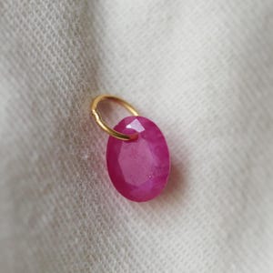 May include: A faceted, oval-shaped, ruby-colored gemstone pendant with a gold-colored loop. The gemstone is a vibrant pinkish-red hue. The pendant is set against a white, textured background.