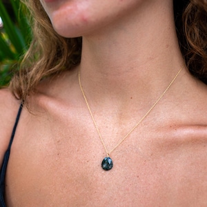 May include: A gold necklace featuring a dark green, teardrop-shaped pendant. The pendant is suspended from a fine gold chain. The necklace is worn against the skin, with a black top visible.