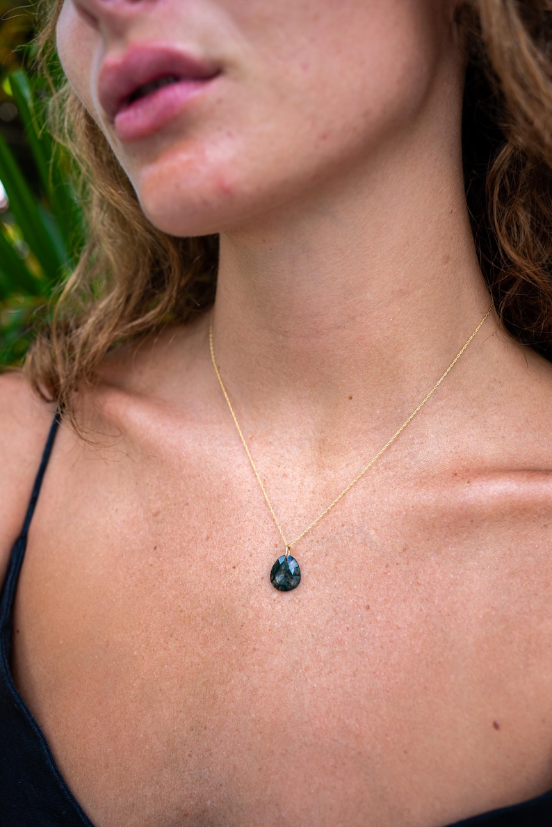 May include: A gold necklace featuring a dark green, teardrop-shaped pendant. The pendant is suspended from a fine gold chain. The necklace is worn against the skin, with a black top visible.