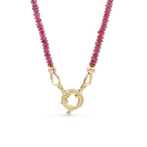 May include: A necklace with a chain of small, reddish-pink gemstones. The necklace is fastened with a gold-coloured clasp, which has a circular design. The clasp is the central feature, with the gemstones forming the necklace's chain.