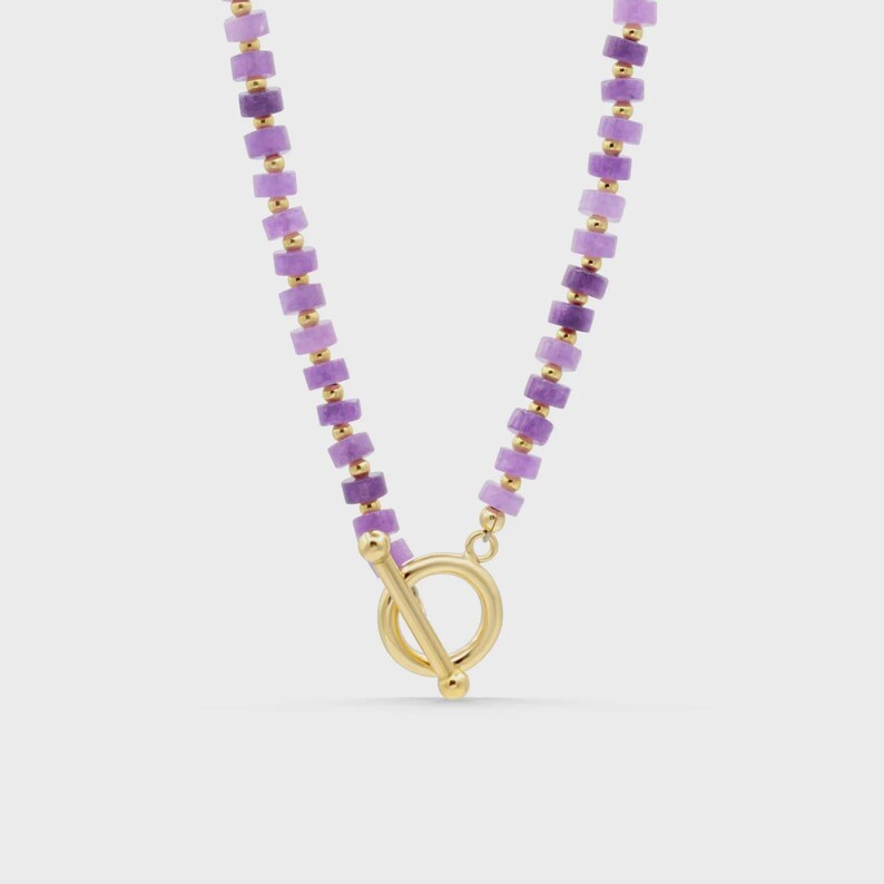 May include: A necklace featuring a series of small, cylindrical purple beads alternating with tiny gold-toned spherical spacers. The piece is secured with a gold-toned toggle clasp consisting of a circular ring and a T-shaped bar closure.
