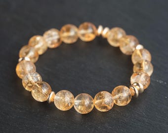 May include: A beaded bracelet featuring translucent, golden-yellow citrine gemstones. The bracelet is strung with small, silver-colored beads between the larger citrine beads. The bracelet is displayed on a dark surface.