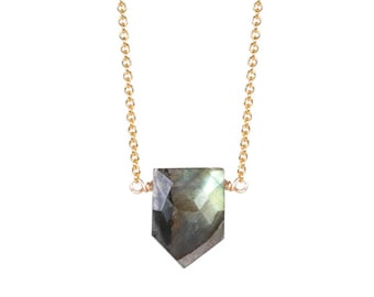 May include: A gold-colored chain necklace with a faceted, shield-shaped labradorite pendant. The pendant is a mix of gray, green, and blue hues. The necklace is displayed against a white background.