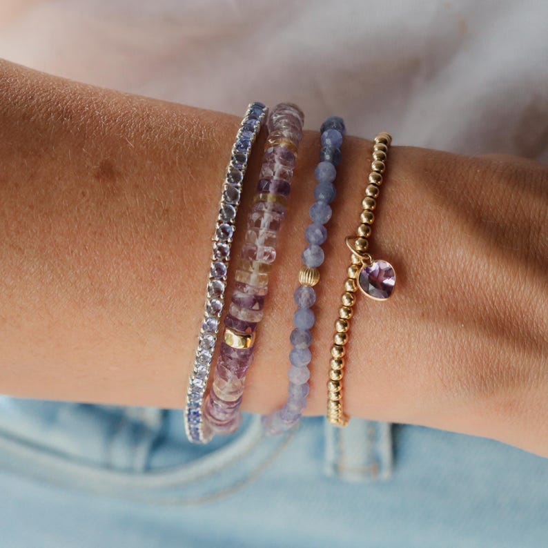 Op de afbeelding: Vier armbanden op een pols. E&eacute;n is een zilveren tennisarmband, de tweede is een mix van paarse en heldere kralen, de derde is blauwe kralen en de vierde is goudkleurig met een paarse hartbedel.