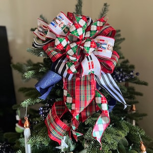 May include: A Christmas tree topper bow with multiple ribbons in red plaid, white with nutcrackers, black and white stripes, and blue glitter. The bow is atop a green Christmas tree with ornaments.