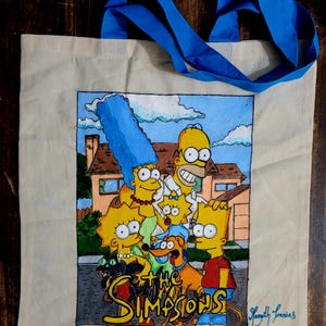 Puede incluir: Una bolsa de lona beige con un asa azul. La parte delantera de la bolsa presenta una imagen pintada a mano de la familia Simpson, incluyendo a Homer, Marge, Bart, Lisa, Maggie, Santa's Little Helper y Snowball. La imagen está situada sobre un fondo de su casa amarilla y un cielo azul con nubes blancas. El texto "The Simpsons" está escrito en amarillo debajo de la imagen. La bolsa está firmada "Katelyn Francis".