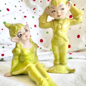 May include: Two ceramic figurines of elves in lime green outfits. One elf is standing with hands near its head, the other is seated. Both have pointed ears, rosy cheeks, and black eyelashes. The background is a white cloth with red and green polka dots.