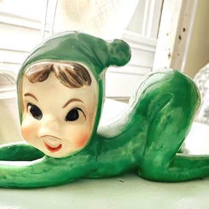 May include: A vintage ceramic figurine of a crawling baby in a green onesie and matching cap. The baby has a fair complexion, rosy cheeks, and brown hair. The figurine is a decorative item.