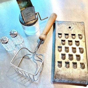 May include: A close-up of a silver grater with a wooden handle, two silver salt shakers, and a vintage nut and meat chopper. The grater has a rectangular shape with a series of holes for grating cheese or other foods. The salt shakers are round and have a small hole at the top for dispensing salt. The nut and meat chopper is a small, hand-cranked device with a blade for chopping nuts or meat.