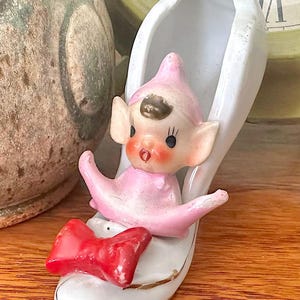 Könnte beinhalten: Eine weiße Keramikfigur in Form eines Schuhs mit einem kleinen, rosa gekleideten Elfen mit spitzen Ohren und brauner Mütze. Der Elf sitzt im Schuh, der mit einer leuchtend roten Schleife an der Spitze und einem Goldrand verziert ist.