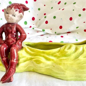 May include: A vintage ceramic planter featuring a seated elf figurine in burgundy attire. The elf is perched on a light green, leaf-shaped planter. The planter has a textured surface and a bright green interior. The background is a white cloth with red and green polka dots.