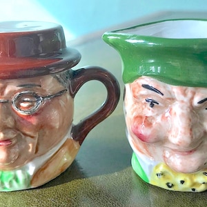 Puede incluir: Dos tazas de té de cerámica con forma de cara. La taza de té de la izquierda lleva un sombrero marrón y gafas. La taza de té de la derecha lleva un sombrero verde. Ambas tazas de té tienen rasgos faciales pintados y sonríen.