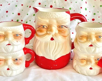 Puede incluir: Una jarra de cerámica de Santa Claus con asa roja y un juego de cuatro tazas a juego de Santa Claus, cada una con expresión de guiño, barba blanca y asa roja, decoradas con adornos florales en tonos rojos y dorados.