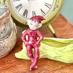Peut inclure: Petite figurine d'elfe en céramique rouge, visage blanc et chapeau pointu. L'elfe est assis sur un plat en céramique vert clair en forme de feuille. Une horloge verte vintage avec des chiffres romains est en arrière-plan.