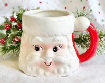 Puede incluir: Una taza de cerámica con forma de cara de Papá Noel con barba, bigote y borde de gorro blancos. La taza presenta un acabado blanco brillante, ojos negros pintados con pestañas, mejillas sonrosadas, boca roja y un asa roja brillante con un pompón blanco.