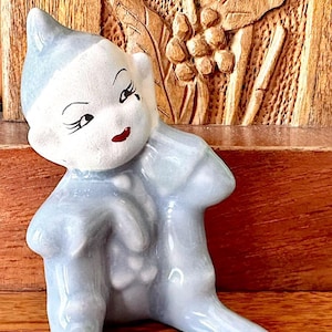 May include: A ceramic figurine of a seated elf with a light blue outfit and a white face. The elf has black eyelashes, a red mouth, and a pointed hat. The background features a wooden carved panel.