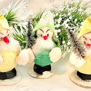 Puede incluir: Tres adornos navideños vintage con forma de muñecos de nieve. Cada muñeco de nieve lleva un sombrero de un color diferente, amarillo, verde y blanco. Los muñecos de nieve sostienen pequeños árboles de cepillo de botella grises.