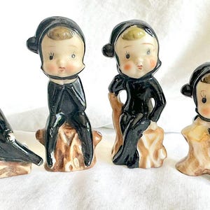May include: Four vintage ceramic figurines of children in black cloaks and hats. Each figure is seated on a brown tree stump. The figurines have rosy cheeks and are in a variety of poses.