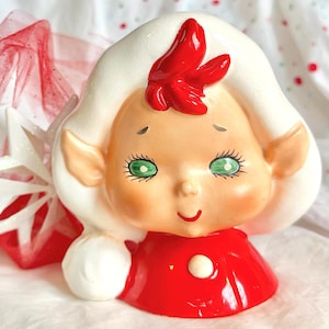 Könnte beinhalten: Eine keramische Weihnachts-Elf-Figur mit weißem Kopf und Haaren, grünen Augen und einer roten Schleife. Der Elf trägt ein rotes Kleid mit einem weißen Knopf. Ein roter und weißer dekorativer Stern befindet sich im Hintergrund.