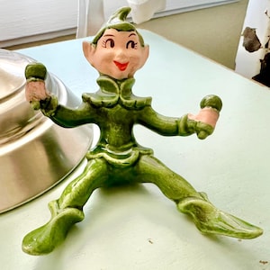 May include: A ceramic figurine of a green elf with a smiling face, rosy cheeks, and a red mouth. The elf is wearing a green outfit and has pink hands. The figurine is posed with arms outstretched.