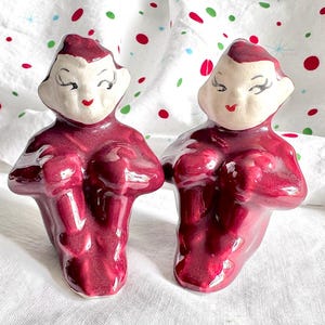 May include: A pair of vintage ceramic figurines. Each figurine is a stylized figure with a burgundy body and a cream-colored face with closed eyes and red lips. The figurines are posed with their hands clasped in front.