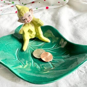 Op de afbeelding: Een vintage keramische blad-vormige schaal in levendig groen, versierd met een kleine gele elf figuur. De schaal bevat drie munten. De elf draagt een gele outfit. Een charmant decoratief stuk.
