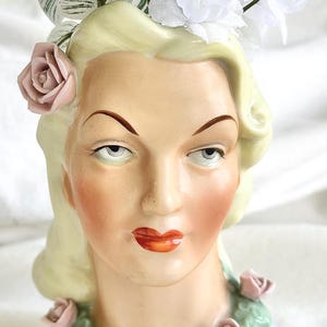 Op de afbeelding: Een vintage keramische hoofdvase in de vorm van een vrouwengezicht, met blond haar en een bleke teint. De vaas is versierd met roze rozen en witte bloemen en heeft een blauwe basis.