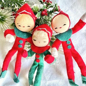 Puede incluir: Tres adornos de duendes navideños vintage. Cada duende tiene una cara redonda y pálida con los ojos cerrados, un gorro puntiagudo rojo y un atuendo rojo o verde. Los duendes tienen cuellos verdes y detalles de botones. Un duende es verde, los otros dos son rojos.