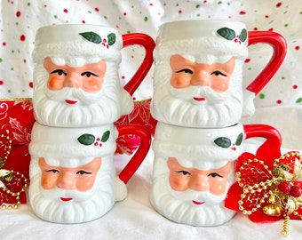 Puede incluir: Conjunto de cuatro tazas de cerámica blanca con forma de Papá Noel con asas rojas. Cada taza presenta una cara de Papá Noel con barba blanca y una rama de acebo verde.