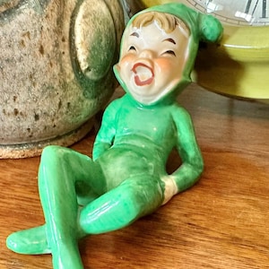 Pode incluir: Estatueta de cerâmica vintage de um elfo sorrindo com roupa e chapéu verdes. O elfo está posando com as pernas cruzadas e luvas brancas. A estatueta tem um rosto pintado com bochechas rosadas e boca aberta. Fundo: relógio.