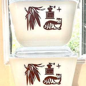 Puede incluir: Dos fuentes de cazuela Pyrex vintage con tapas de vidrio transparente. Los platos blancos presentan ilustraciones marrones de maíz, un molinillo de café, un gato y una cruz. Los platos están apilados.