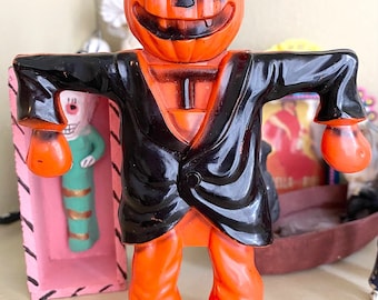 Könnte beinhalten: Eine Vintage-Halloween-Dekoration eines Kürbis-Kopf-Vogelscheuchens mit einem schwarzen Anzug und orangefarbenen Hosen. Die Vogelscheuche ist aus Plastik und hat ein freundliches, zahniges Grinsen.