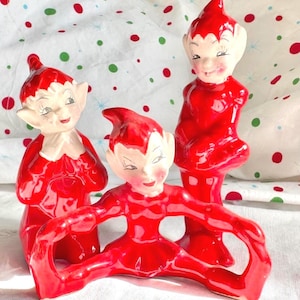 May include: Three ceramic elf figurines with glossy red outfits and pointed hats. Two elves are standing in upright positions, while the third elf is positioned in the front, performing a split with arms extended outward to the sides.