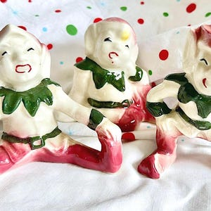Könnte beinhalten: Drei Keramik-Elfenfiguren mit weißer Haut, spitzen Ohren und roten Mützen. Jeder Elf trägt einen grünen Kragen und Gürtel sowie eine rote Hose und Stiefel. Die Elfen sind in verschiedenen sitzenden Positionen mit ausdrucksstarken Gesichtern dargestellt.