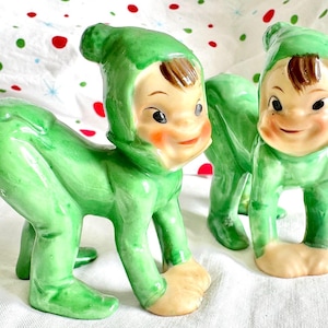 Peut inclure: Deux figurines vintage en céramique représentant une créature enfantine en combinaison et capuche vertes. Les figurines ont un visage souriant avec des joues roses, des cheveux bruns et des pieds bronzés. Les figurines sont en position de ramper.