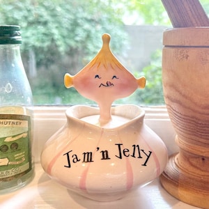 Op de afbeelding: Een witte keramische pot met roze strepen en een glimlachend cartoon gezicht op de deksel. De pot heeft de tekst "Jam 'n Jelly" in zwarte letters.