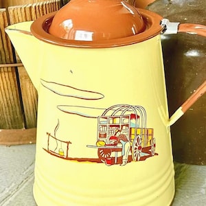 Op de afbeelding: Gele emaille koffiepot met een bruin deksel en rand. De pot heeft een gedetailleerde illustratie van een huifkar en een persoon die boven een vuur kookt. De handgreep is bruin en de tuit bevindt zich aan de linkerkant.
