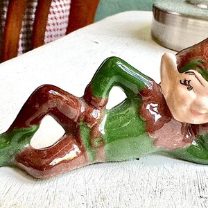 Puede incluir: Una figura de elfo de cerámica en posición reclinada. El elfo está pintado con un cuerpo verde, piernas marrones y un sombrero marrón y verde. La cara del elfo es de color melocotón claro con los ojos cerrados.