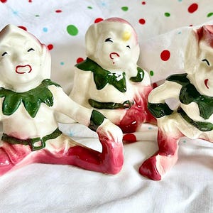 Puede incluir: Tres figuras de elfos de cerámica con trajes rojos y blancos, cuellos verdes y caras sonrientes. Los elfos están en varias poses, con uno sentado y dos arrodillados. Las figuras están sobre una superficie blanca con lunares rojos y verdes.