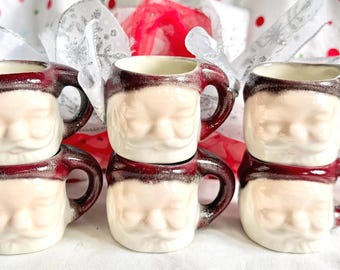 Puede incluir: Un juego de seis tazas de cerámica con forma de caras de Papá Noel, cada una con un rostro color crema con bigote y un gorro rojo oscuro con acabado texturizado. Las tazas están dispuestas en dos columnas verticales de tres unidades.