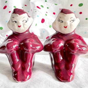 May include: Pair of vintage ceramic figurines in a deep burgundy color. The figurines have a stylized design with a light beige face, red lips, and closed eyes. The figures are holding their hands in front of them, with a glossy finish.
