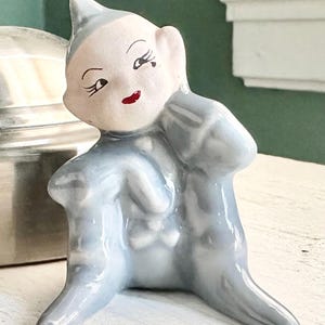 May include: A vintage ceramic figurine of a seated elf with a pointed hat. The elf has a pale face with red lips and black eyelashes. The figurine is primarily light blue with a glossy finish, and is posed with one arm raised.