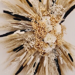 May include: A dried floral arrangement with white and cream flowers, pampas grass, and other dried grasses. The arrangement is in a burst shape and is mounted on a white wall.