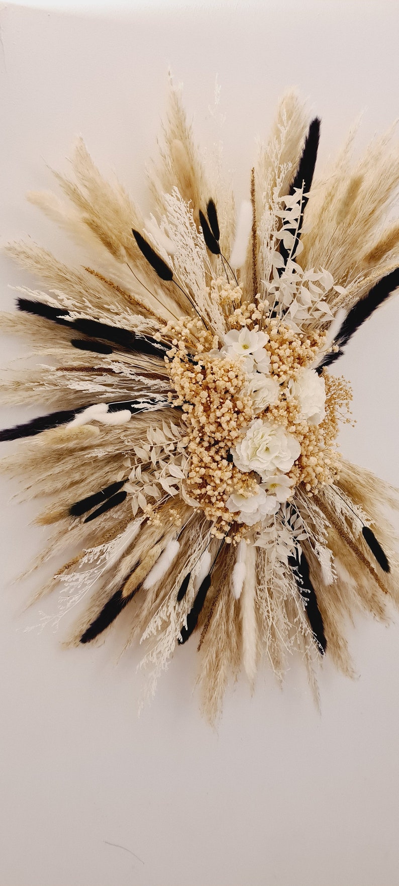 May include: A dried floral arrangement with white and cream flowers, pampas grass, and other dried grasses. The arrangement is in a burst shape and is mounted on a white wall.
