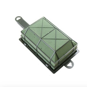 May include: A green floral foam brick with a black plastic frame and handle. The foam is divided into sections with a grid pattern.