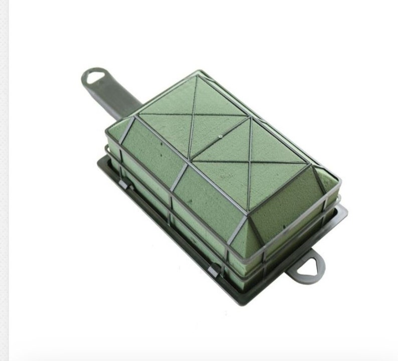 May include: A green floral foam brick with a black plastic frame and handle. The foam is divided into sections with a grid pattern.