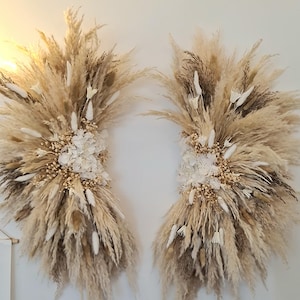 May include: Two large, cream-coloured dried flower arrangements with pampas grass, dried hydrangeas, and other dried flowers. The arrangements are hanging on a white wall.